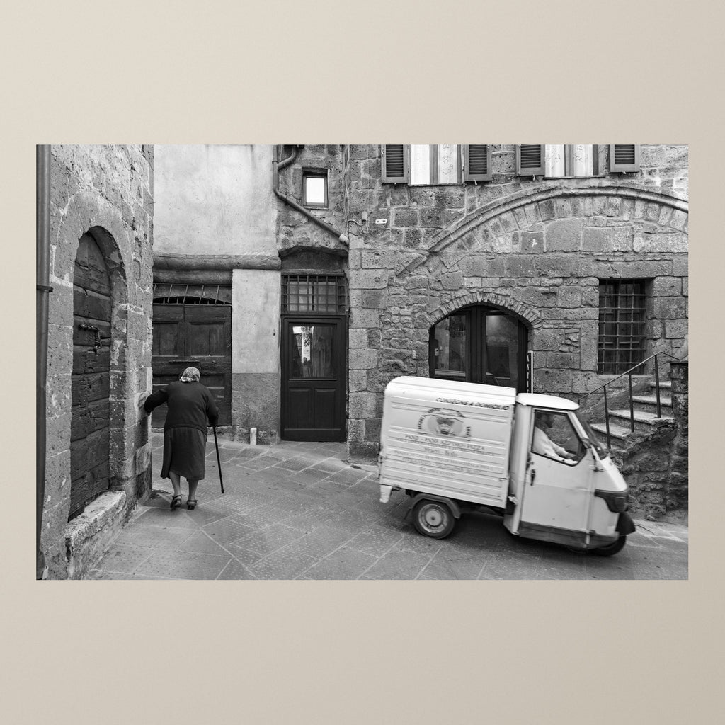 Everyday Eternity – A Piaggio Ape by ancient stone walls as an elderly woman walks by – photograph by Duccio Nacci