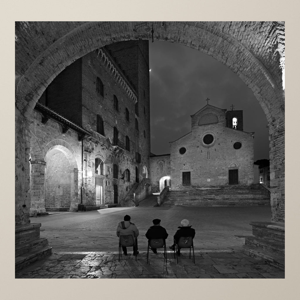 In the Cathedral's Shadow – Three elderly people seated on chairs facing the façade of a Gothic church – photograph by Duccio Nacci