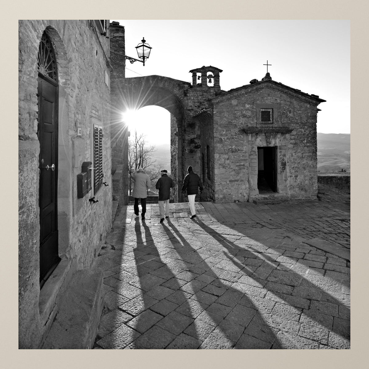 Night's Shadow – Three figures walk beneath a stone arch as the setting sun casts long shadows – photograph by Duccio Nacci