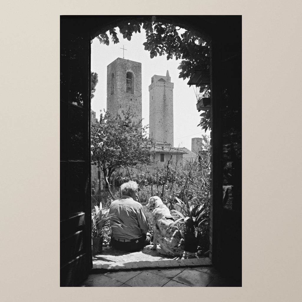Silent Guardians – A man and his dog in a garden under a stone arch, the towers of San Gimignano behind – photograph by Duccio Nacci