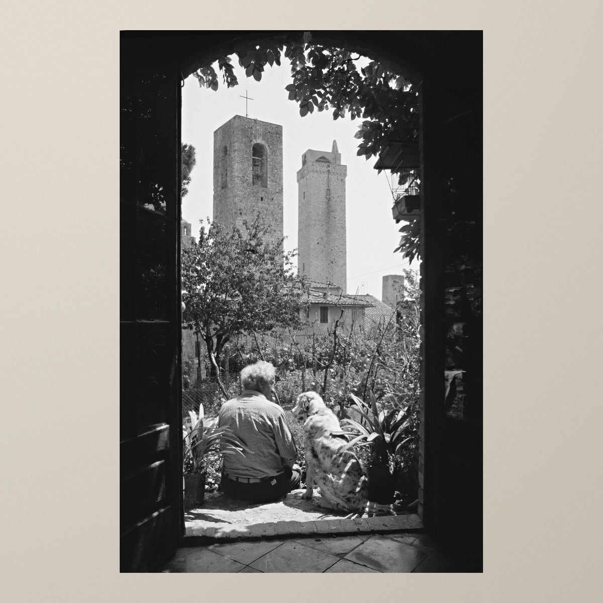 Silent Guardians – A man and his dog in a garden under a stone arch, the towers of San Gimignano behind – photograph by Duccio Nacci