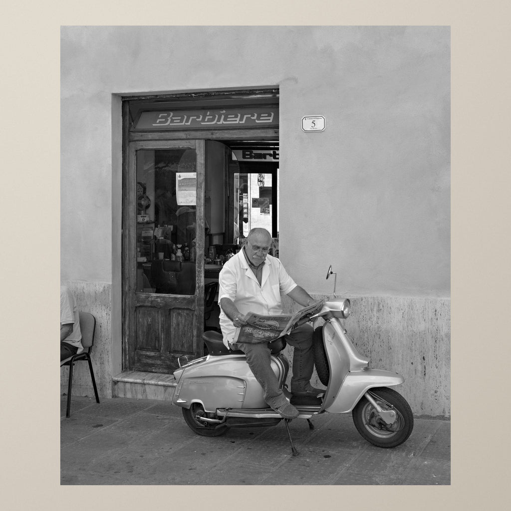 The Barber on the Vespa – A barber reads the newspaper sitting on his Vespa outside his shop – photograph by Duccio Nacci