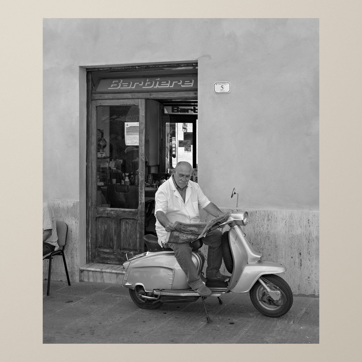 The Barber on the Vespa – A barber reads the newspaper sitting on his Vespa outside his shop – photograph by Duccio Nacci