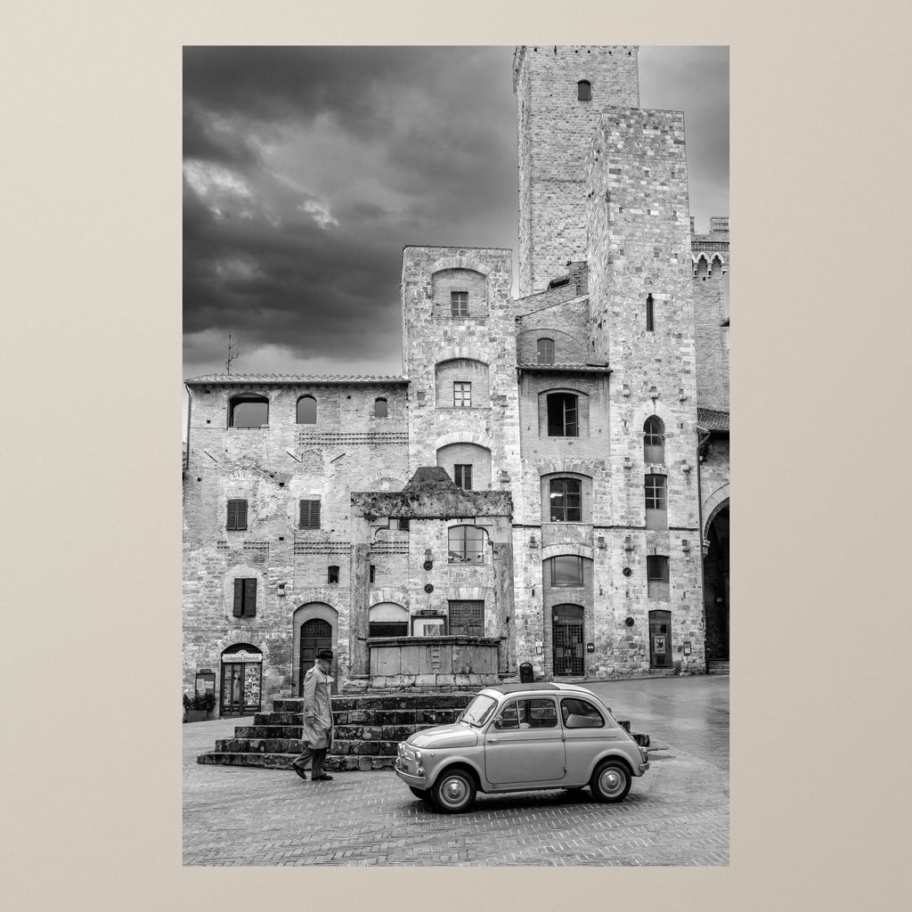 The Gentleman Stroll – A Fiat 500 before the towers of San Gimignano as a gentleman in a trenchcoat walks by – photograph by Duccio Nacci