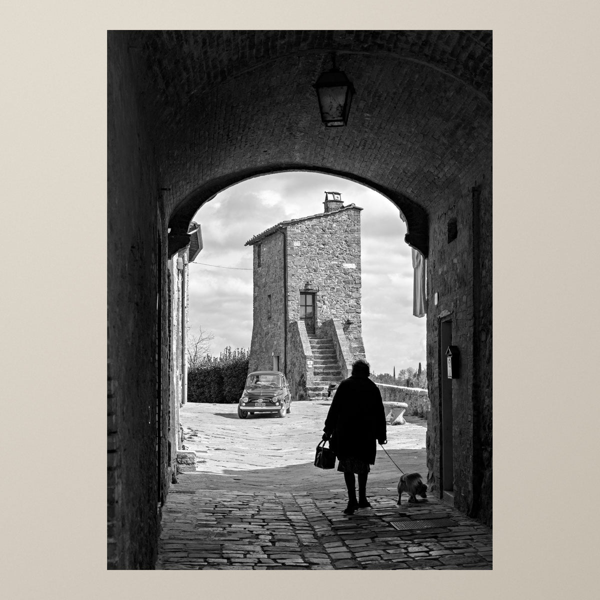 Through the Archway – An elderly lady walks her dog under a medieval arch, a Fiat 500 parked behind – photograph by Duccio Nacci